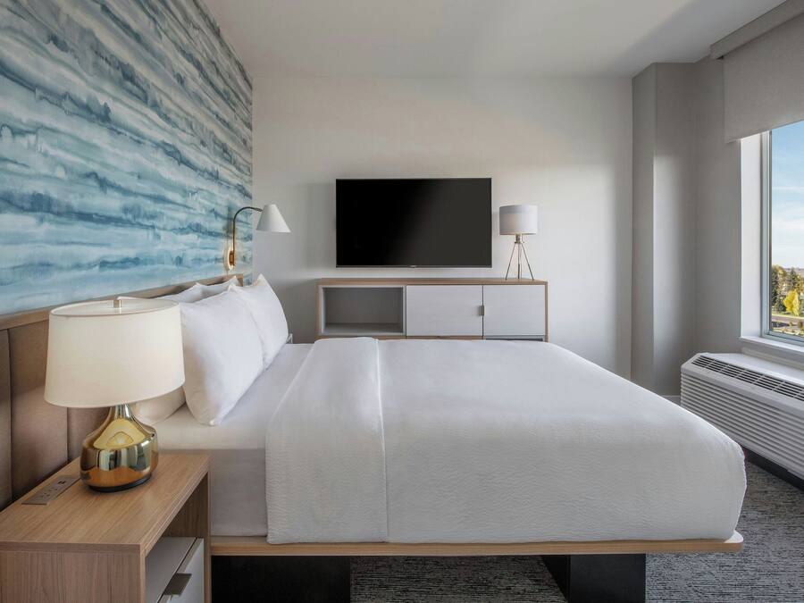 Moderni hotellihuone, jossa on suuri sänky, taulutelevisio, yöpöytälamppu ja ikkunasta avautuva näkymä kaupunkiin.