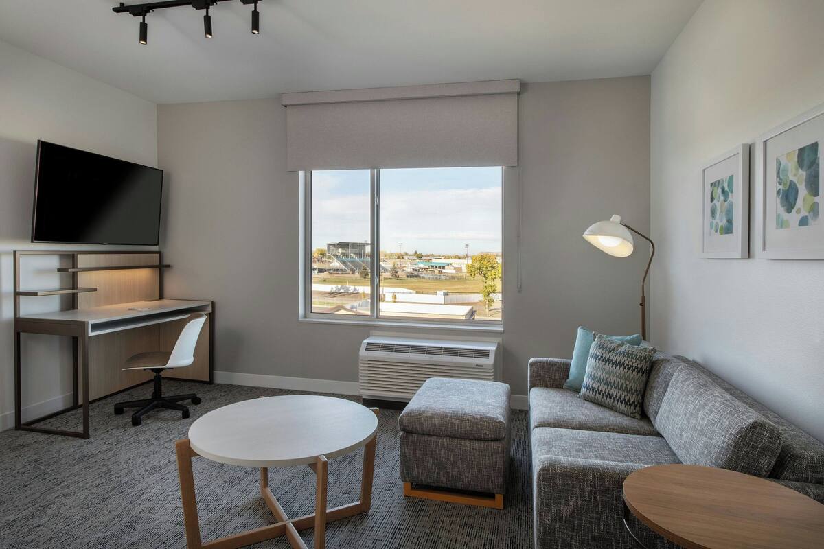 Une chambre d’hôtel moderne équipée d’un téléviseur à écran plat, d’un bureau avec une chaise, d’une table ronde, d’un canapé et d’une grande fenêtre donnant sur l’extérieur.
