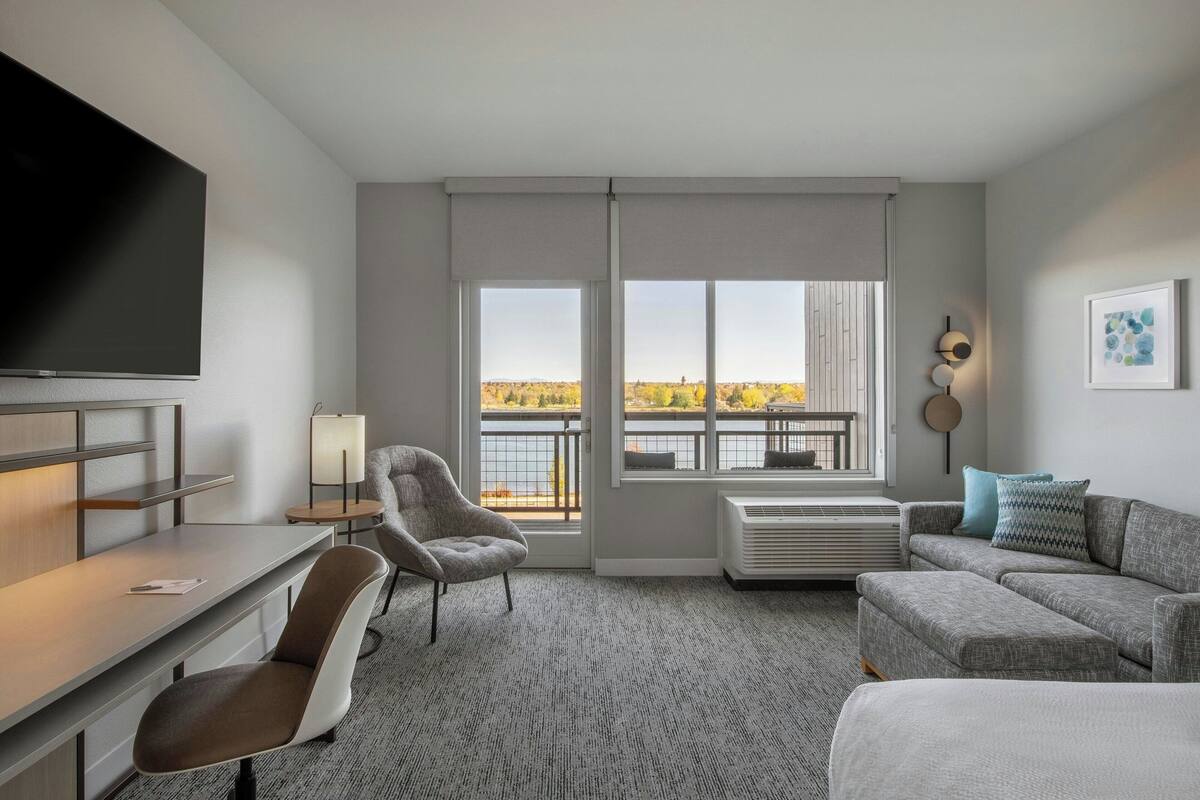 Une chambre d’hôtel moderne dotée d’une télévision à écran plat, d’un bureau, d’un fauteuil confortable, d’un canapé et de grandes fenêtres offrant une vue sur un plan d’eau et le feuillage automnal.