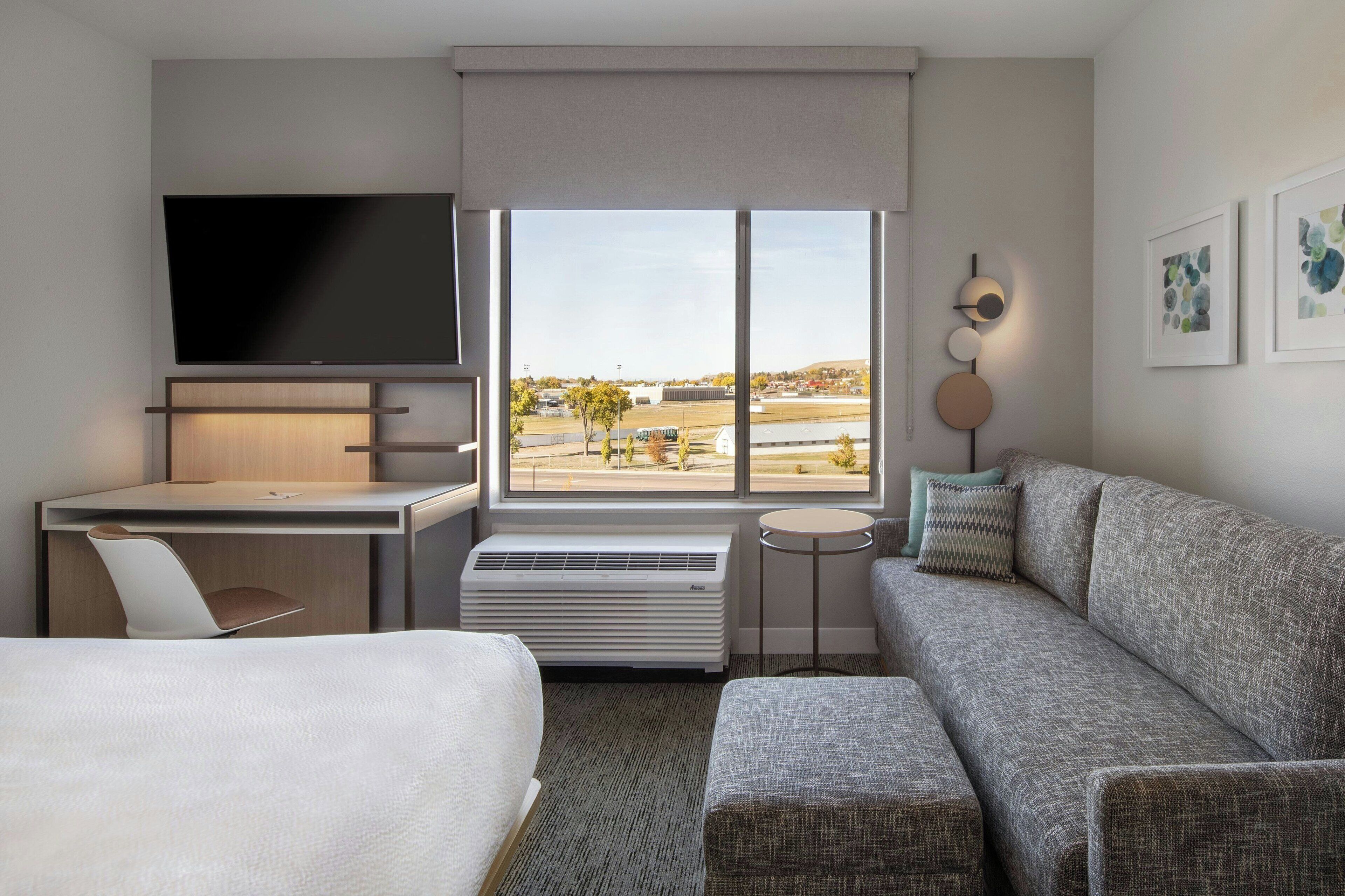 A hotel room with a bed, a desk with a chair, a sofa, a TV, a large window with a view, and a small table.