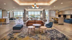 A modern lounge area with a sofa, round tables, and chairs, large windows offering a view, and a kitchen area in the background. - Towneplace Suites By Marriott Great Falls (Great Falls)