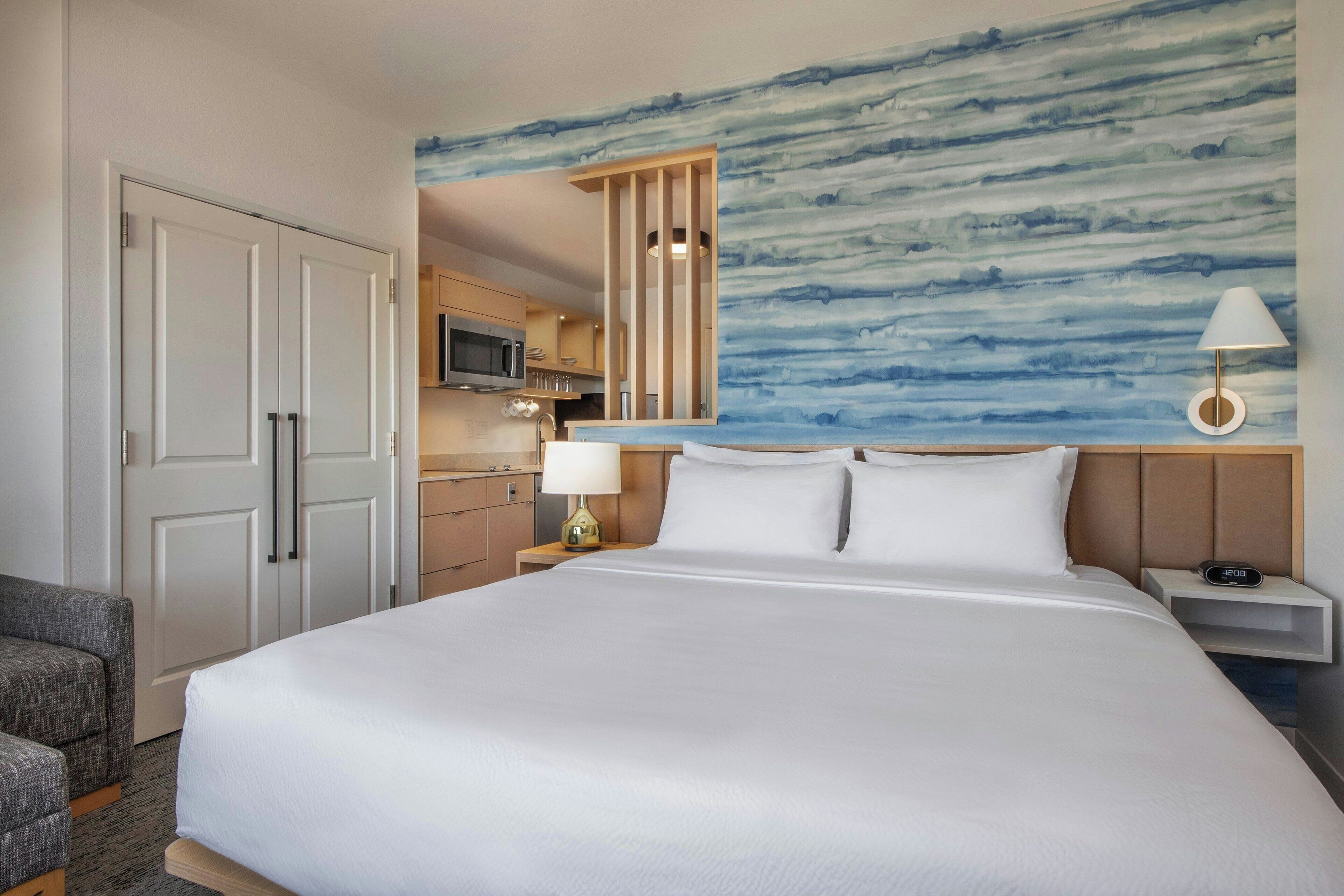 A hotel room with a large bed, a desk with a lamp, a TV, and a kitchenette.