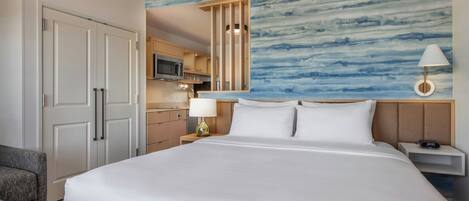 A hotel room with a large bed, a desk with a lamp, a TV, and a kitchenette.