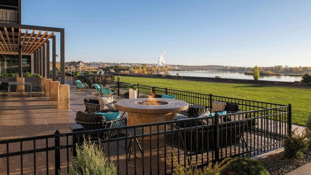An outdoor patio with a fire pit, seating, and a view of a lake and distant buildings. - Towneplace Suites By Marriott Great Falls (Great Falls)