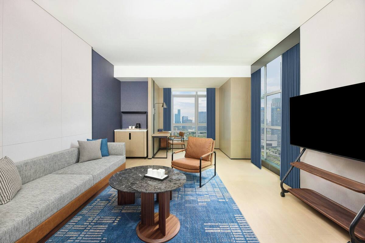 Una habitación de hotel moderna con sofá, una mesita de centro redonda, televisión, zona de comedor con mesa y sillas, y vistas a la ciudad a través de amplios ventanales.