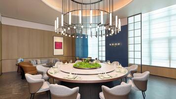 A modern dining area with a round table set for six, surrounded by six chairs. A unique chandelier hangs above the table, and the room features a large window with a view.