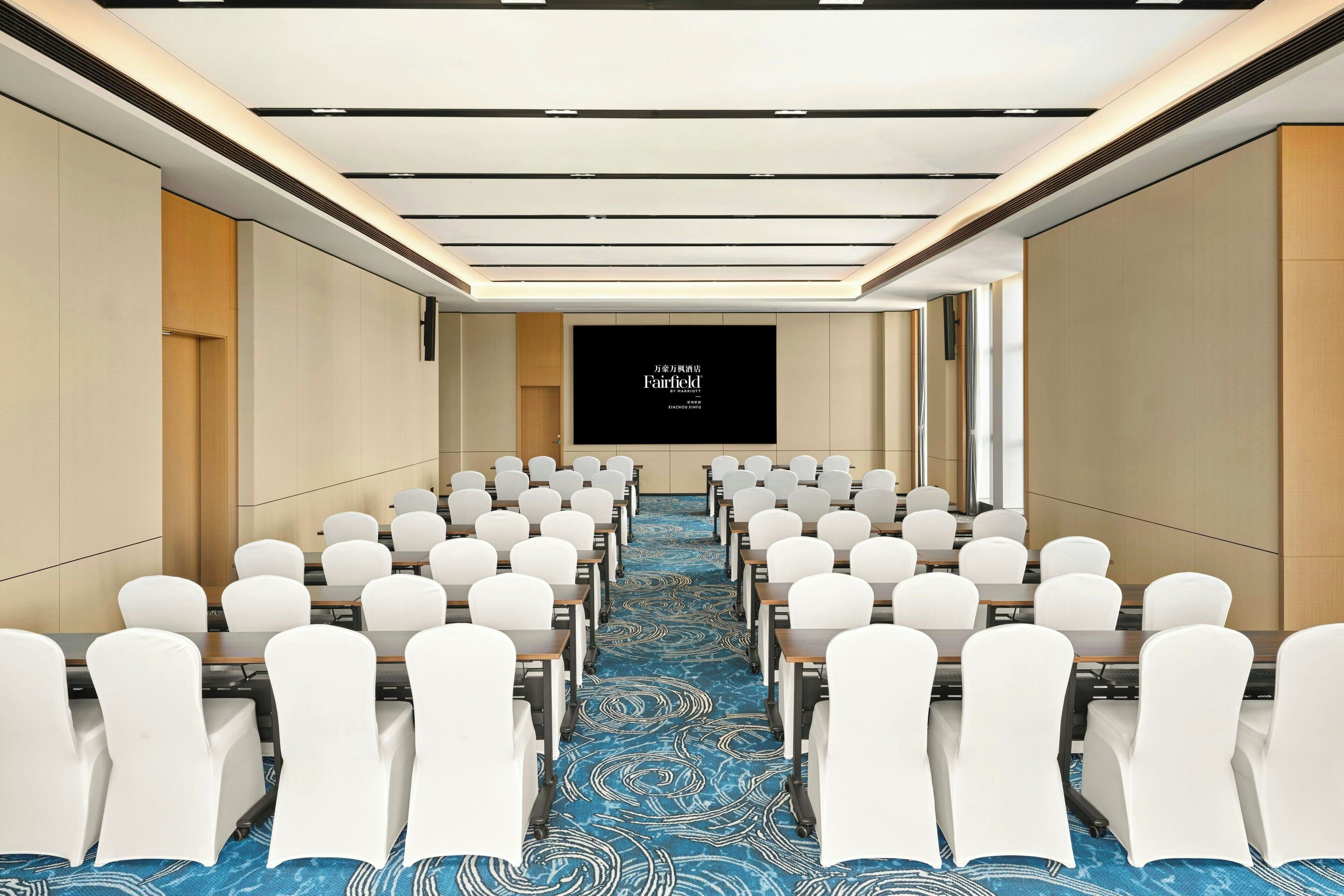 A conference room with rows of white chairs, a blue patterned carpet, and a large screen at the front.