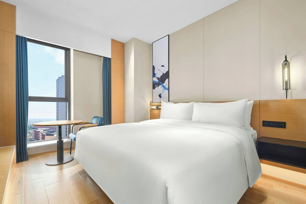 A modern hotel room with a large bed, a small table, and a view of a cityscape through a large window.