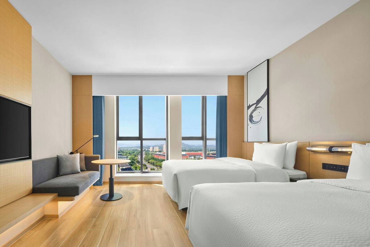 A hotel room with a large window, two beds, a sofa, a desk, and a flat-screen TV.