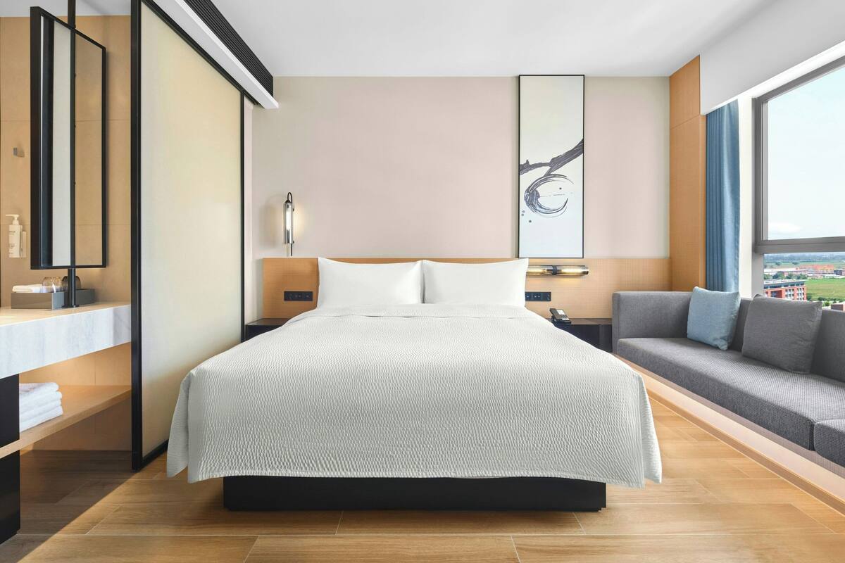 A modern hotel room with a large bed, a sofa, a bedside table, a mirror, and a window with a city view.