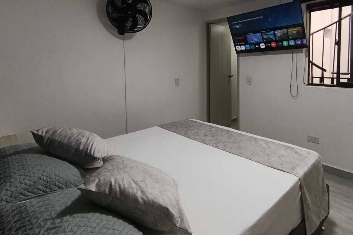 A bedroom with a bed, pillows, a standing fan, a control panel, and a window with bars.