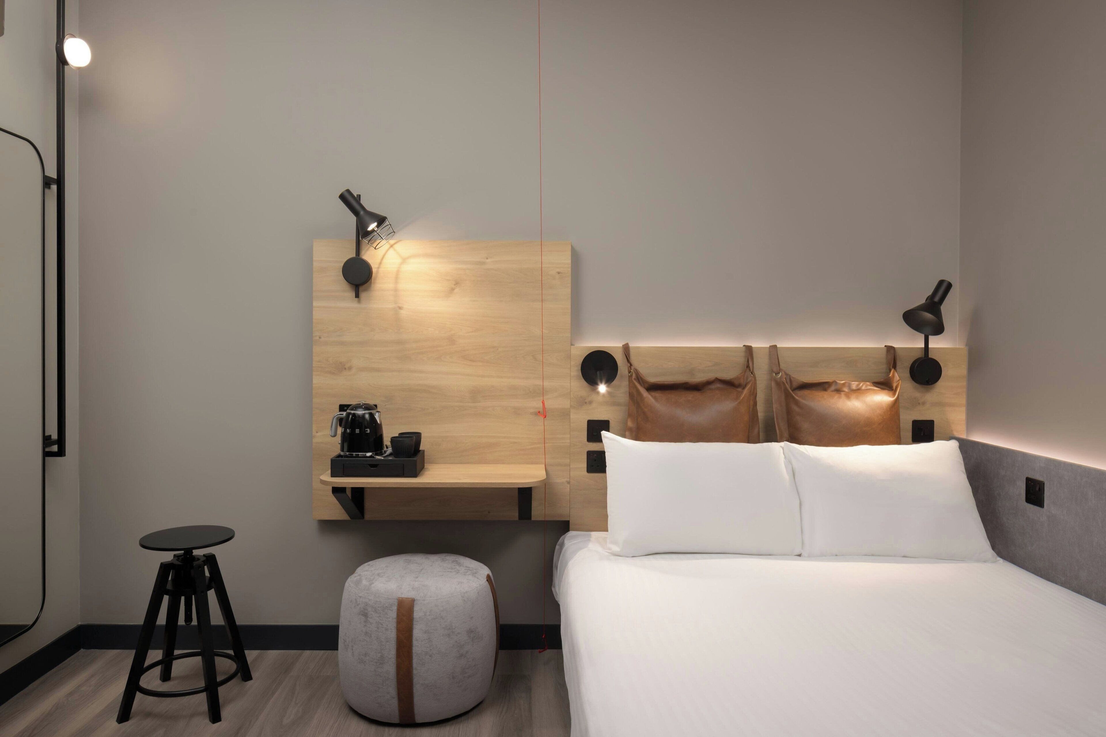 A modern hotel room with a wooden headboard, a small desk with a coffee machine, a round ottoman, and a bed with white linens.