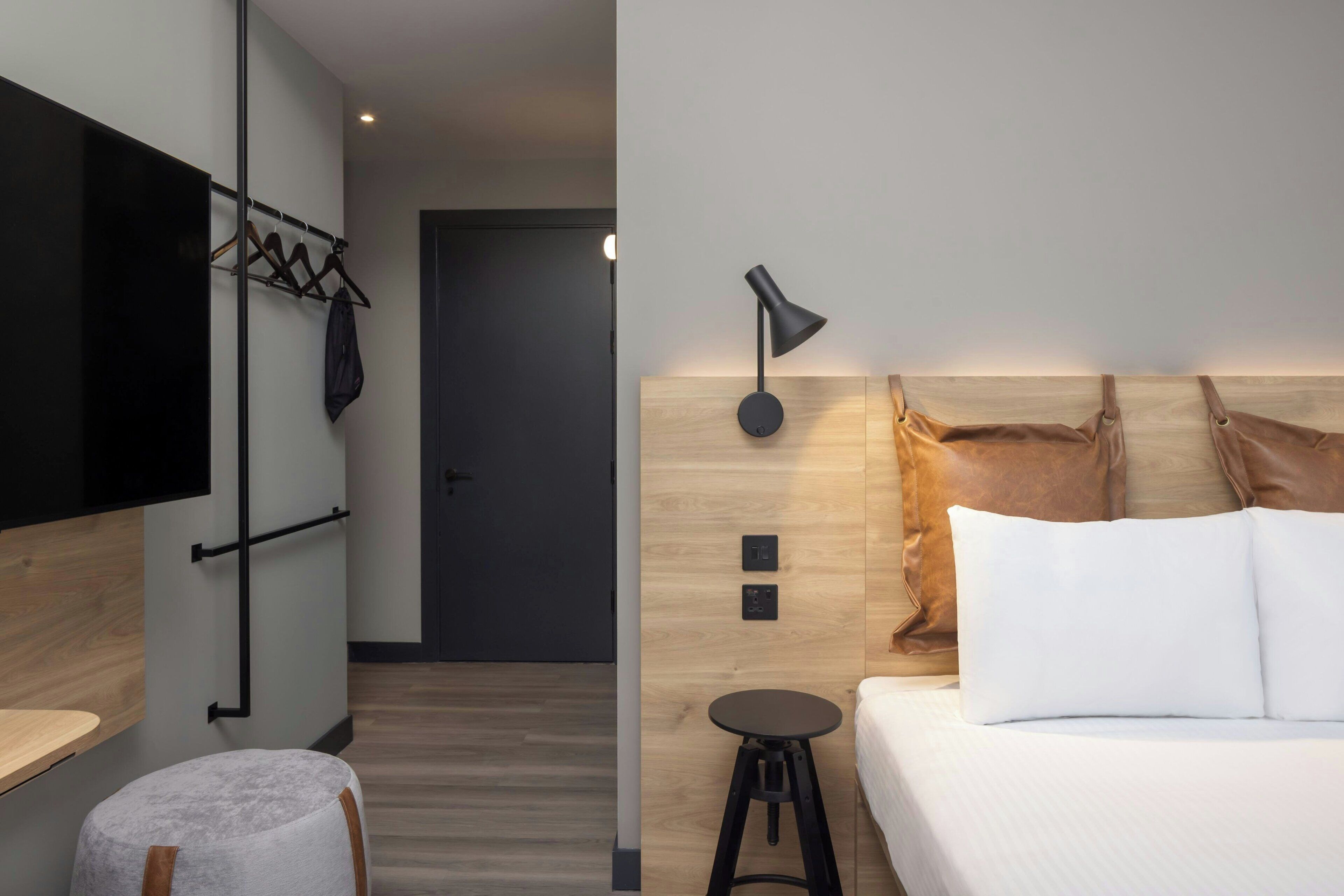 A modern hotel room with a bed, a small stool, a flat-screen TV mounted on the wall, a coat rack, and a wooden headboard with pillows.