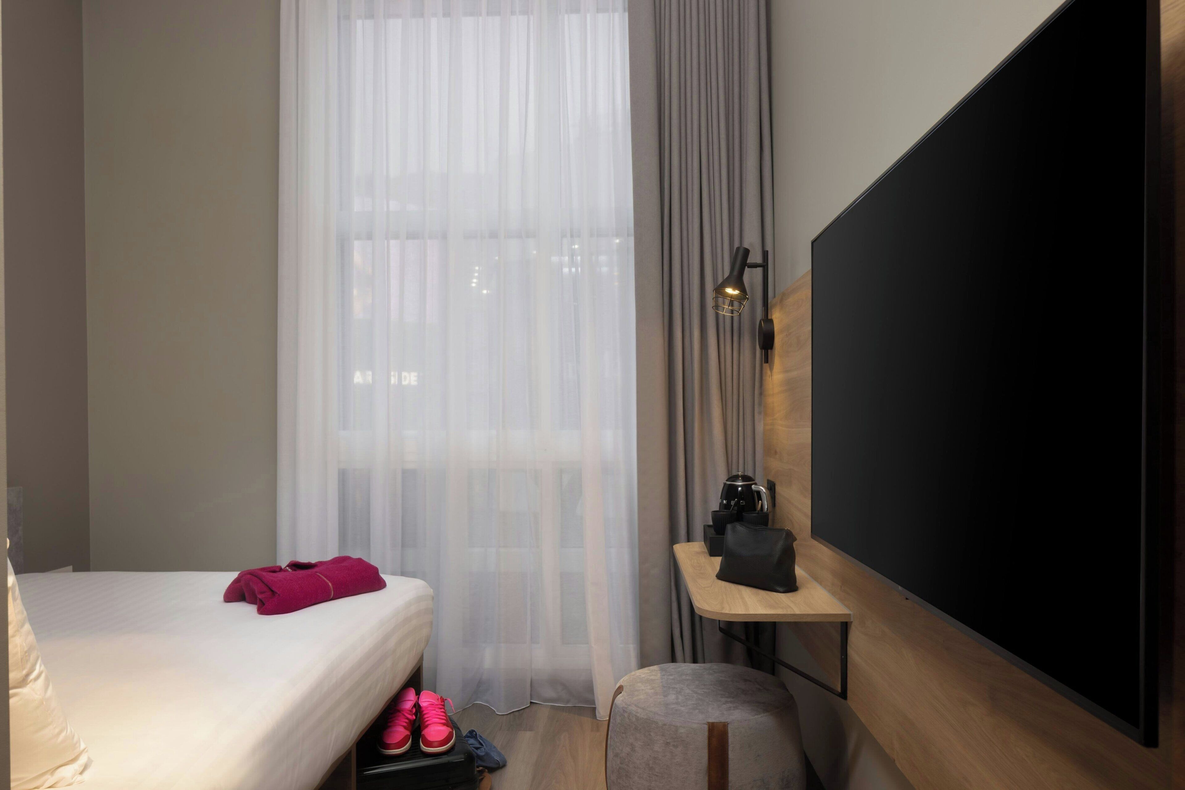 A hotel room with a bed, a TV, a small table with a coffee maker, and a window with sheer curtains.