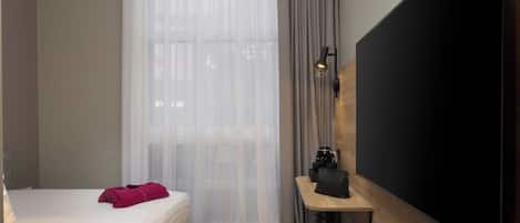 A hotel room with a bed, a TV, a small table with a coffee maker, and a window with sheer curtains.