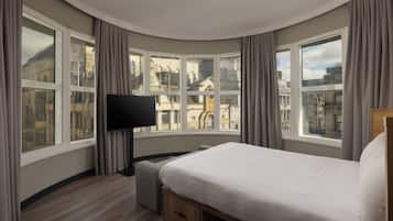 Habitación de hotel con una cama, un televisor de pantalla plana, una mesita y vistas a edificios de la ciudad a través de amplias ventanas.