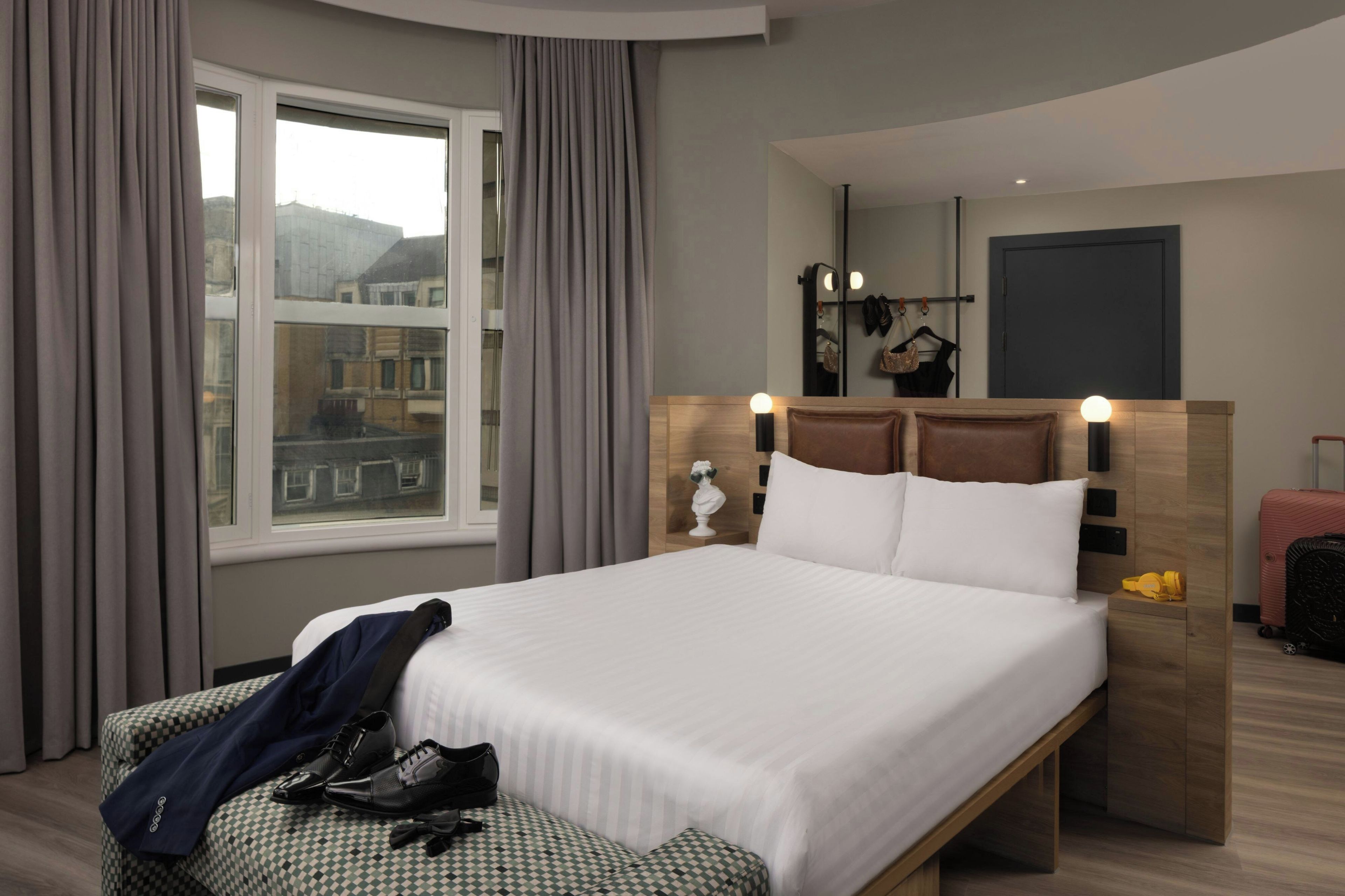 A hotel room with a large bed, a wooden headboard, a bedside table with a lamp, a mirror, and a view of buildings outside the window.