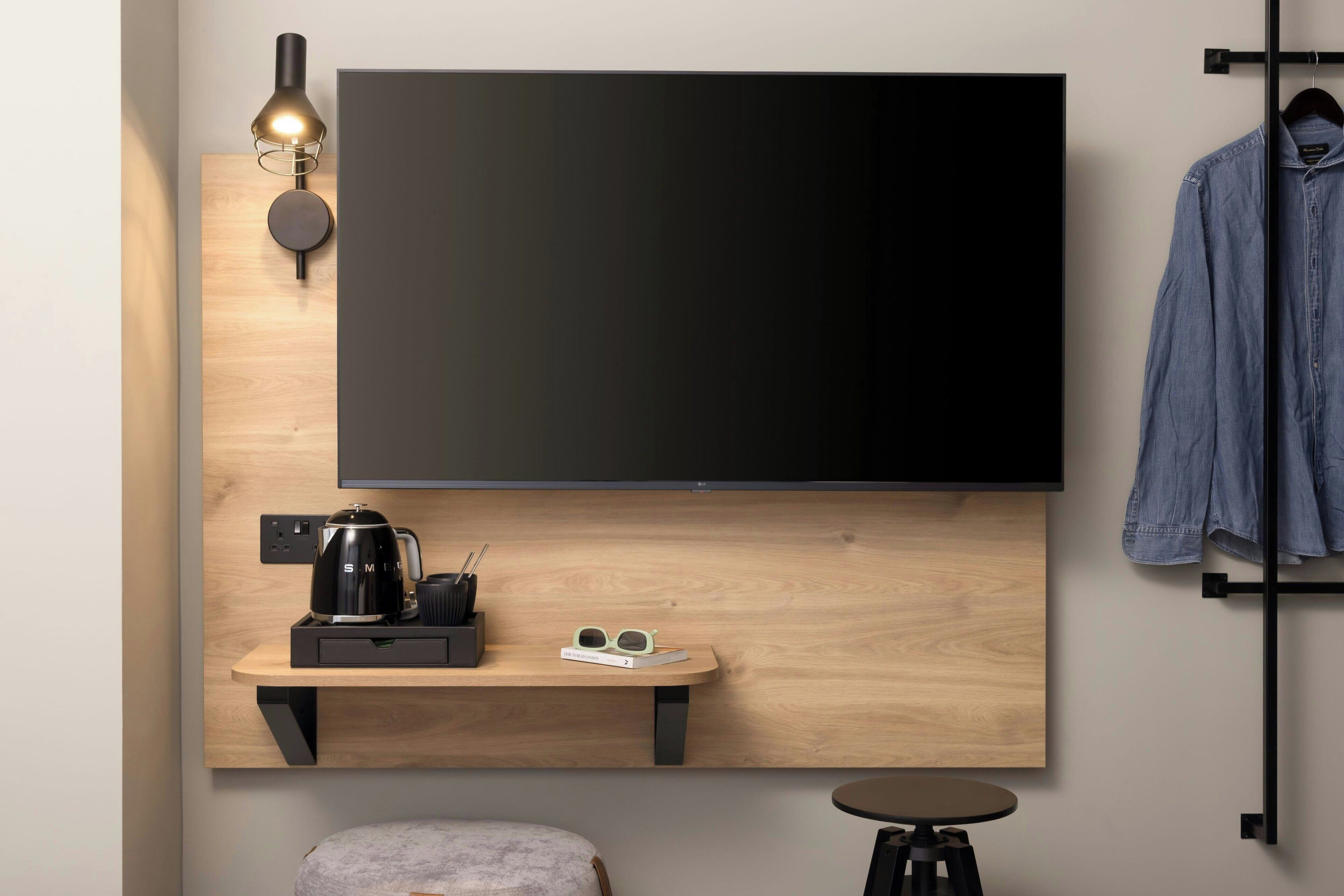 A modern room with a large flat-screen TV mounted on a wooden wall, a small wooden desk with a coffee maker, glasses, and a remote control, a round ottoman, and a hanging rack with a denim jacket.
