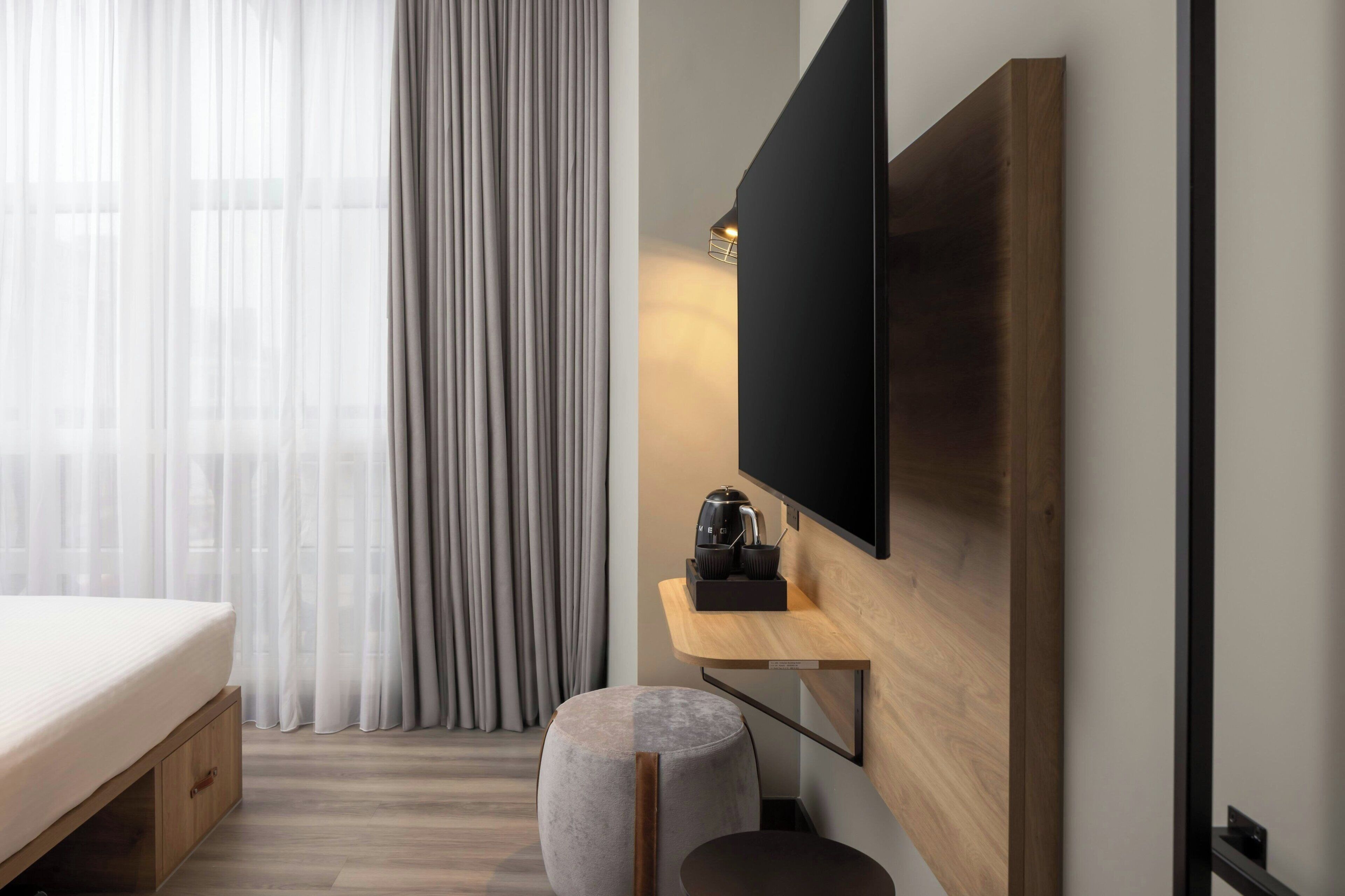 Ett modernt hotellrum med en säng, en stor platt-TV som är monterad på väggen, ett träbord som sängbord, en liten rund pall och tunna, genomskinliga gardiner.