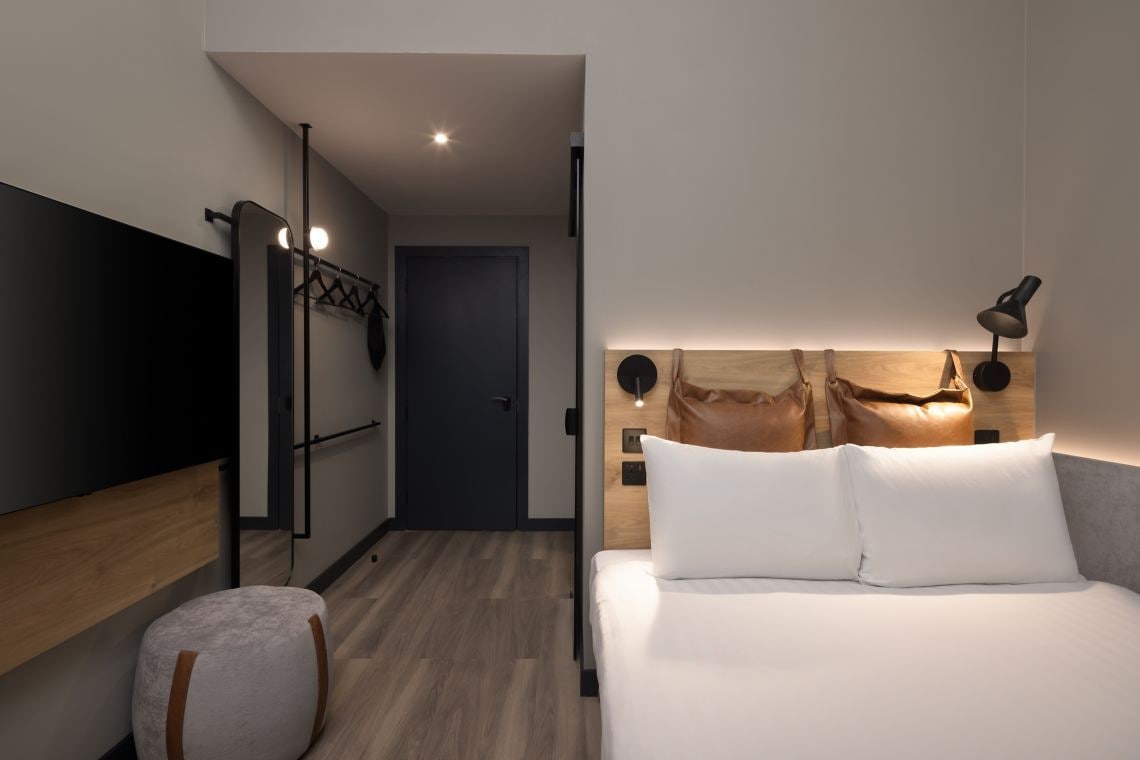 Une chambre d’hôtel moderne équipée d’un lit, d’une lampe de chevet, d’une tête de lit en bois et d’un téléviseur fixé au mur.