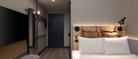 Une chambre d’hôtel moderne équipée d’un lit, d’une lampe de chevet, d’une tête de lit en bois et d’un téléviseur fixé au mur.