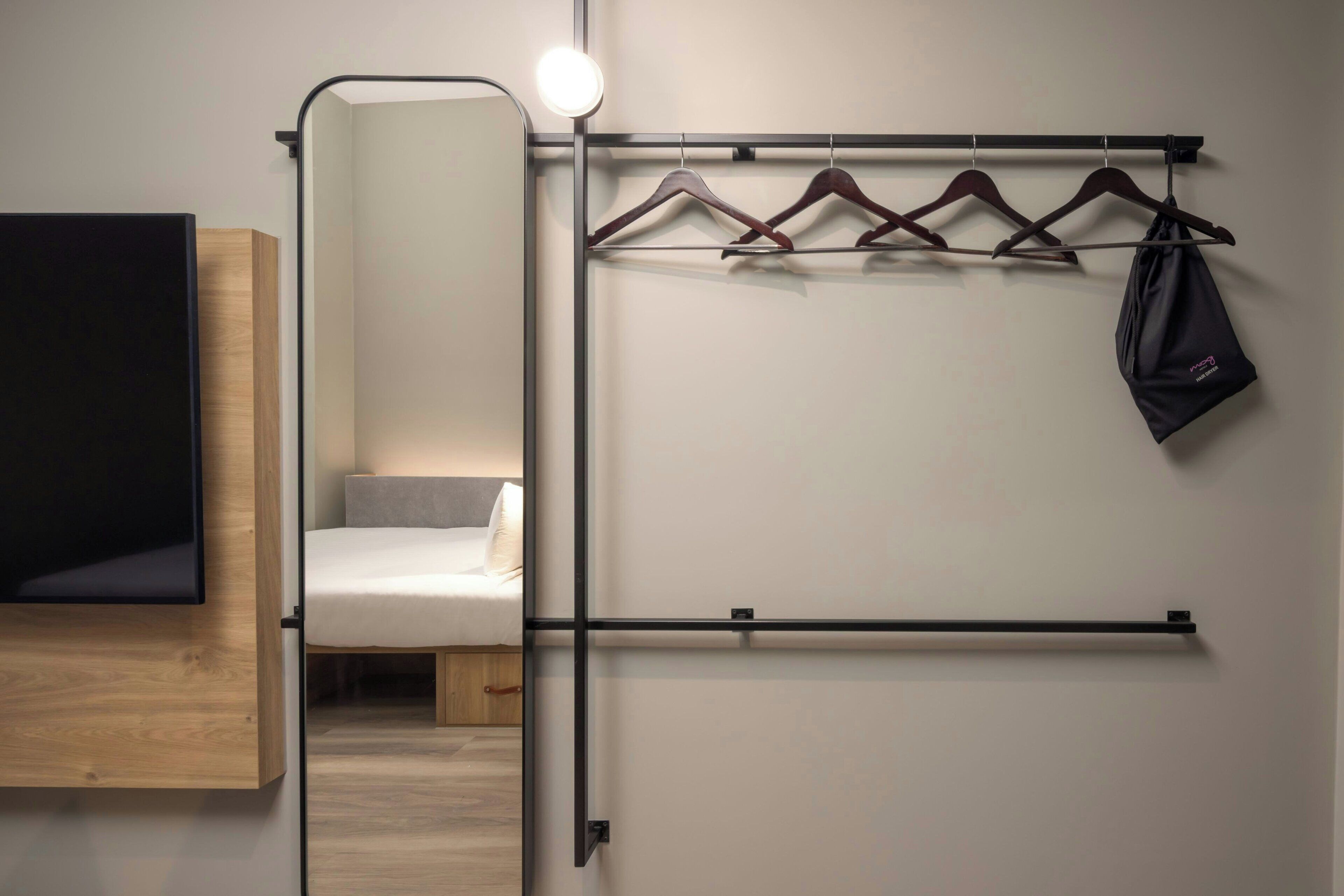 Une chambre moderne équipée d’un lit, d’un miroir, d’un porte-manteau avec des cintres et d’un sac noir.