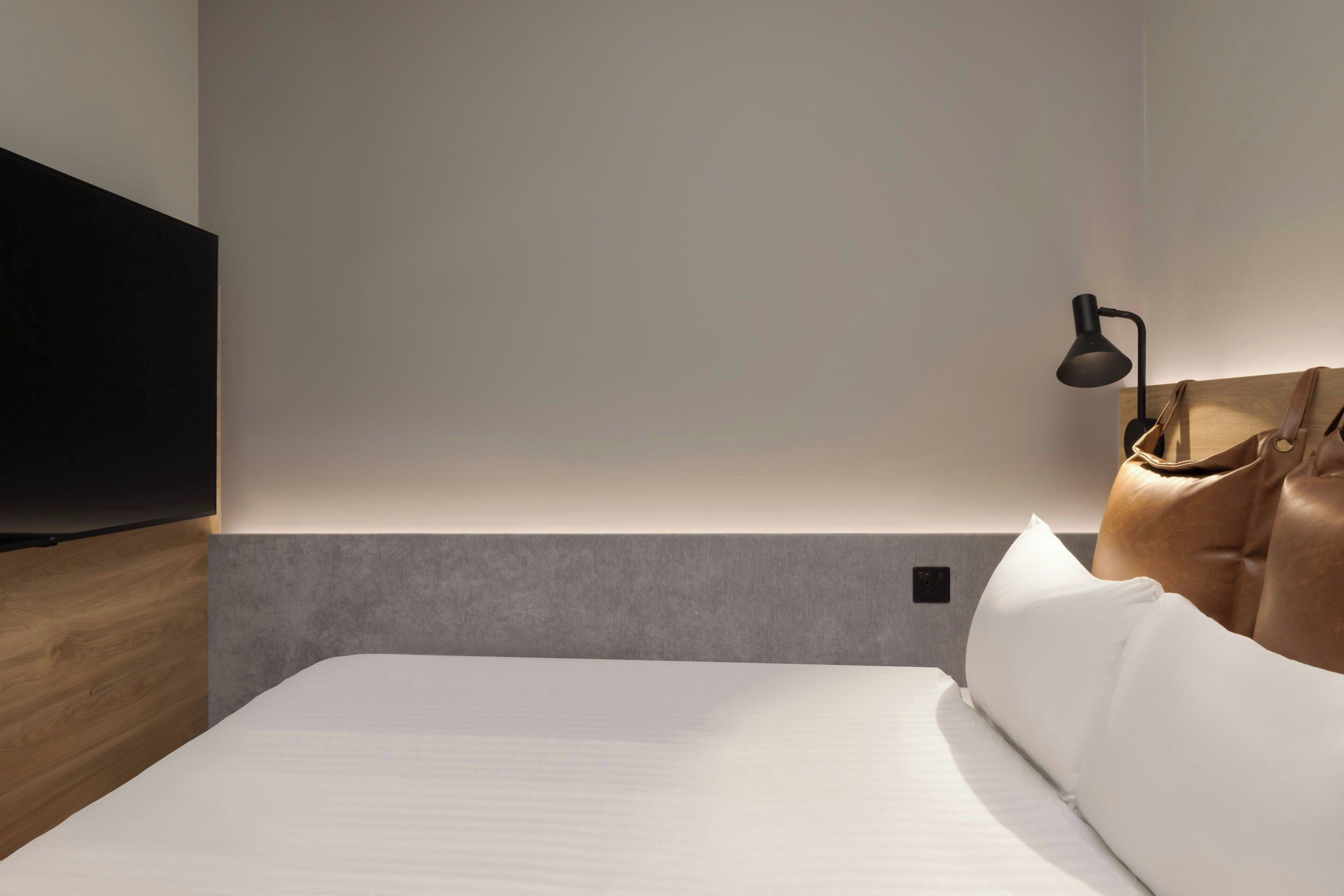A hotel room with a bed, a flat-screen TV mounted on the wall, a bedside lamp, and a leather bag on the bed.