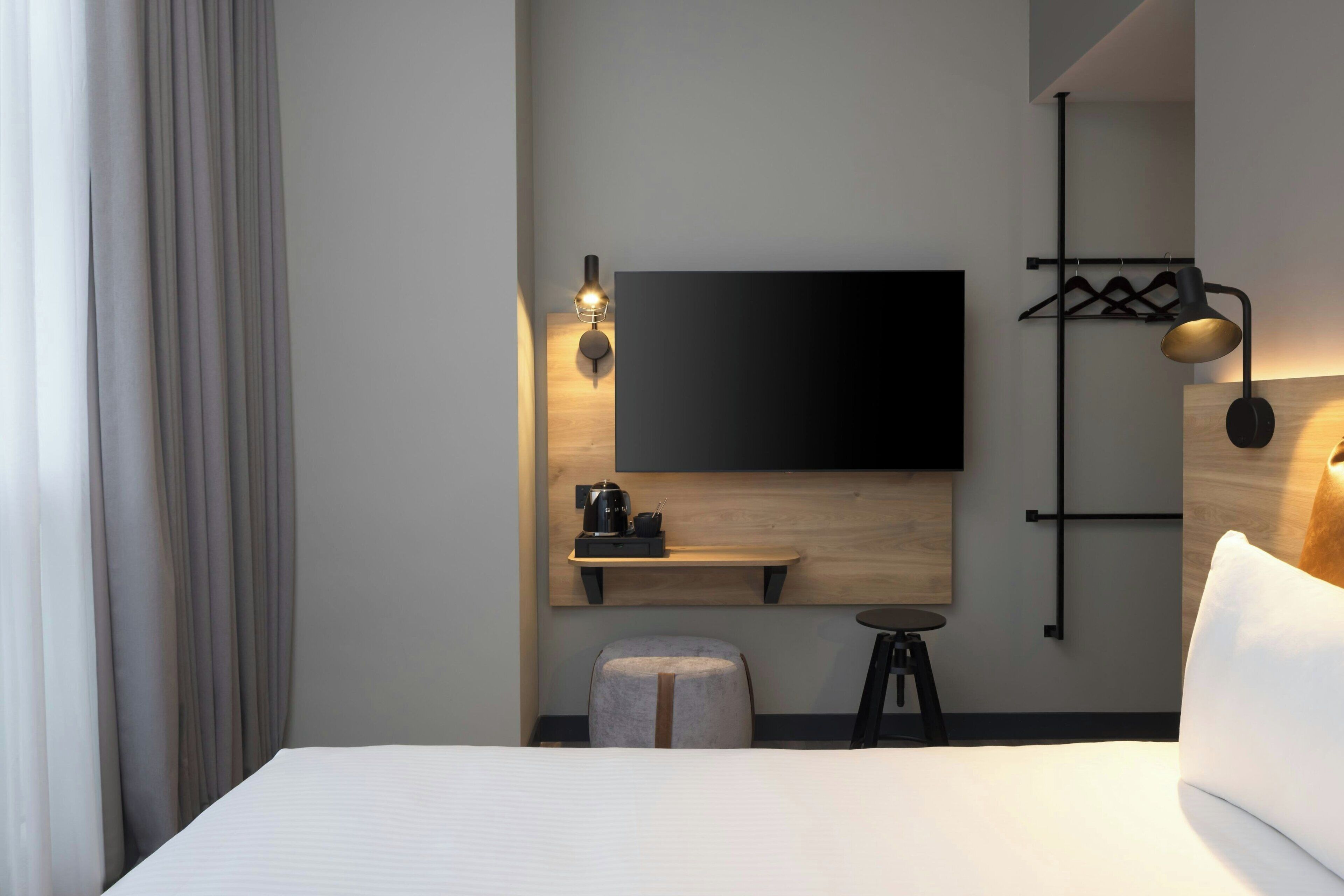 A modern hotel room with a large bed, a flat-screen TV mounted on the wall, a small table with a kettle, and a stool.