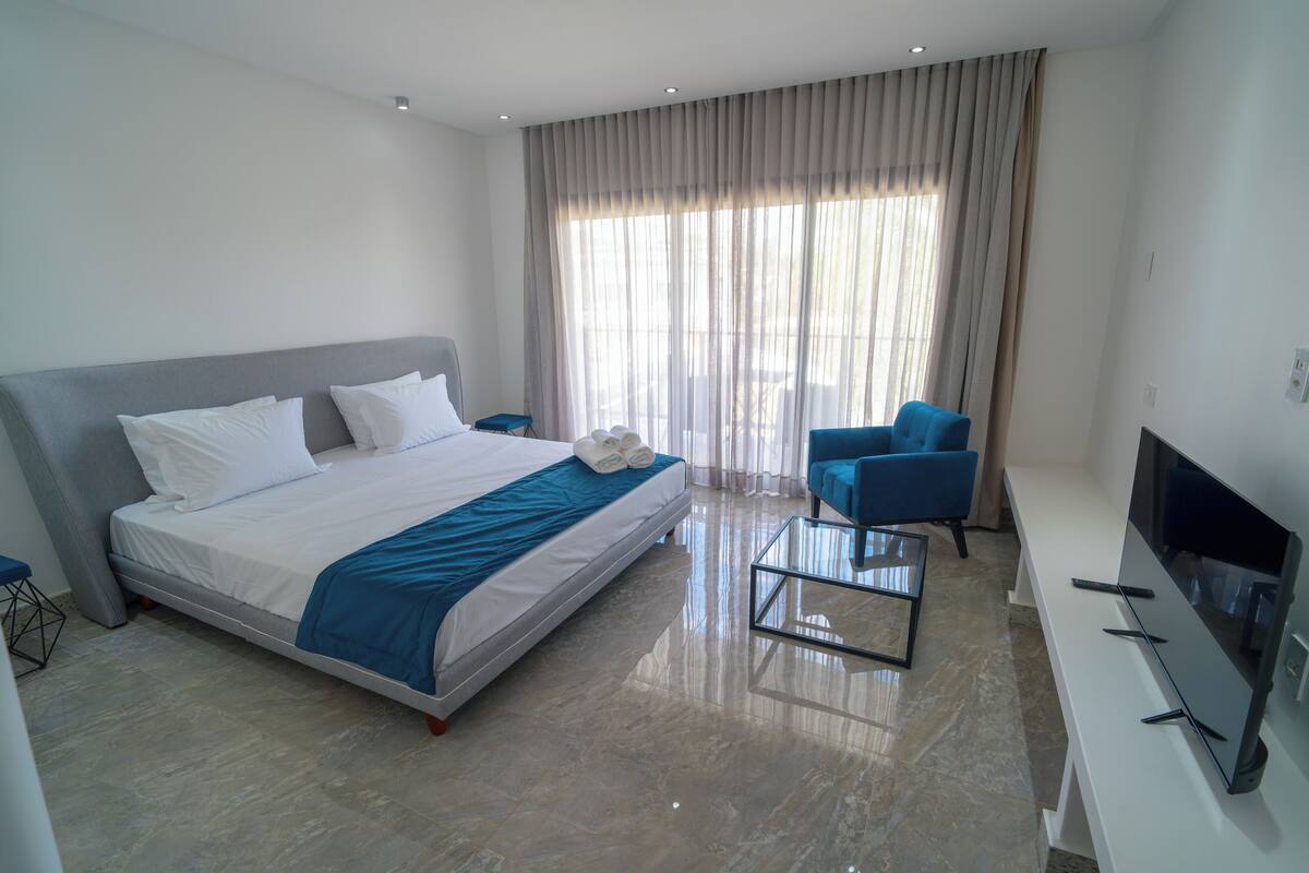 A modern hotel room with a large bed, a TV, a seating area, and a view of the outdoors.