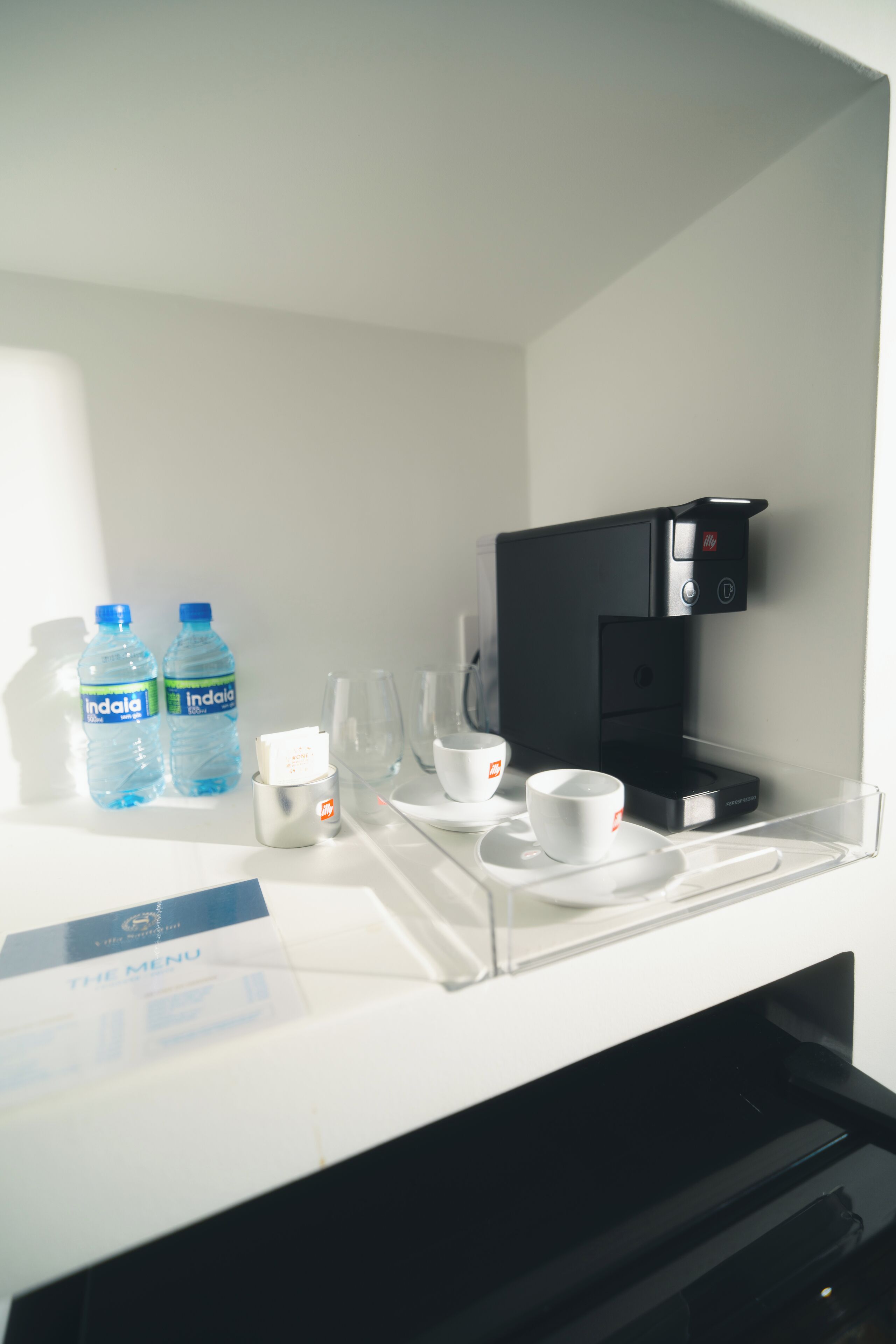 Eine Minibar im Hotel mit einer Kaffeemaschine, zwei Wasserflaschen und einer Speisekarte.