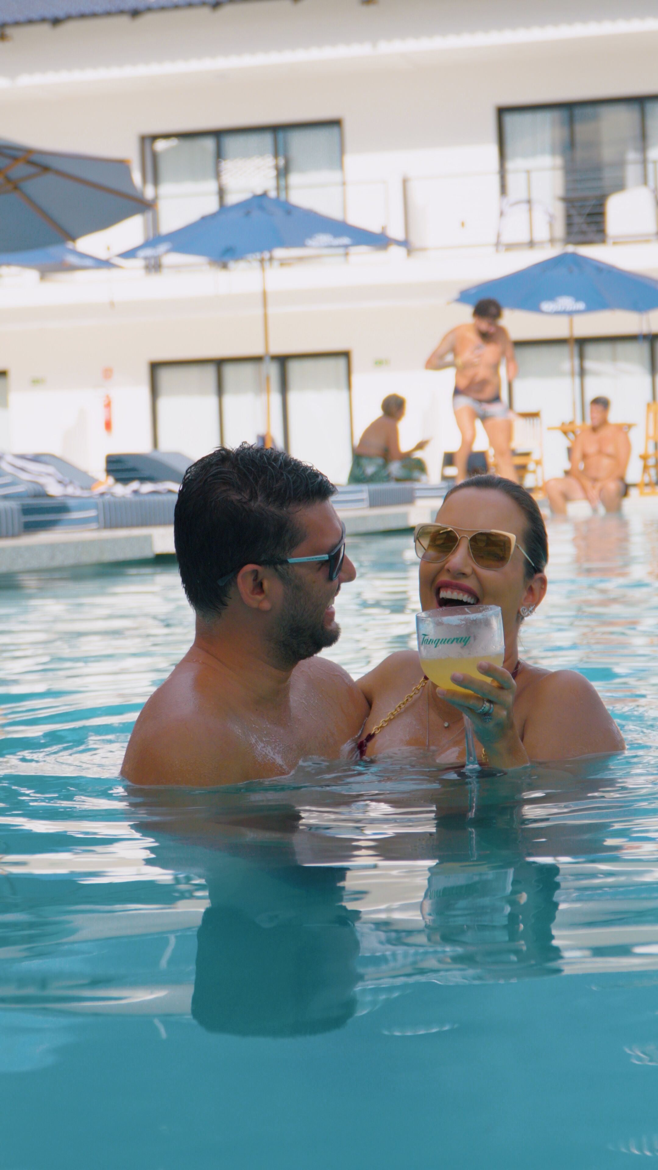 Um casal em uma piscina, com vista clara para a área da piscina, incluindo guarda-sóis e espreguiçadeiras.