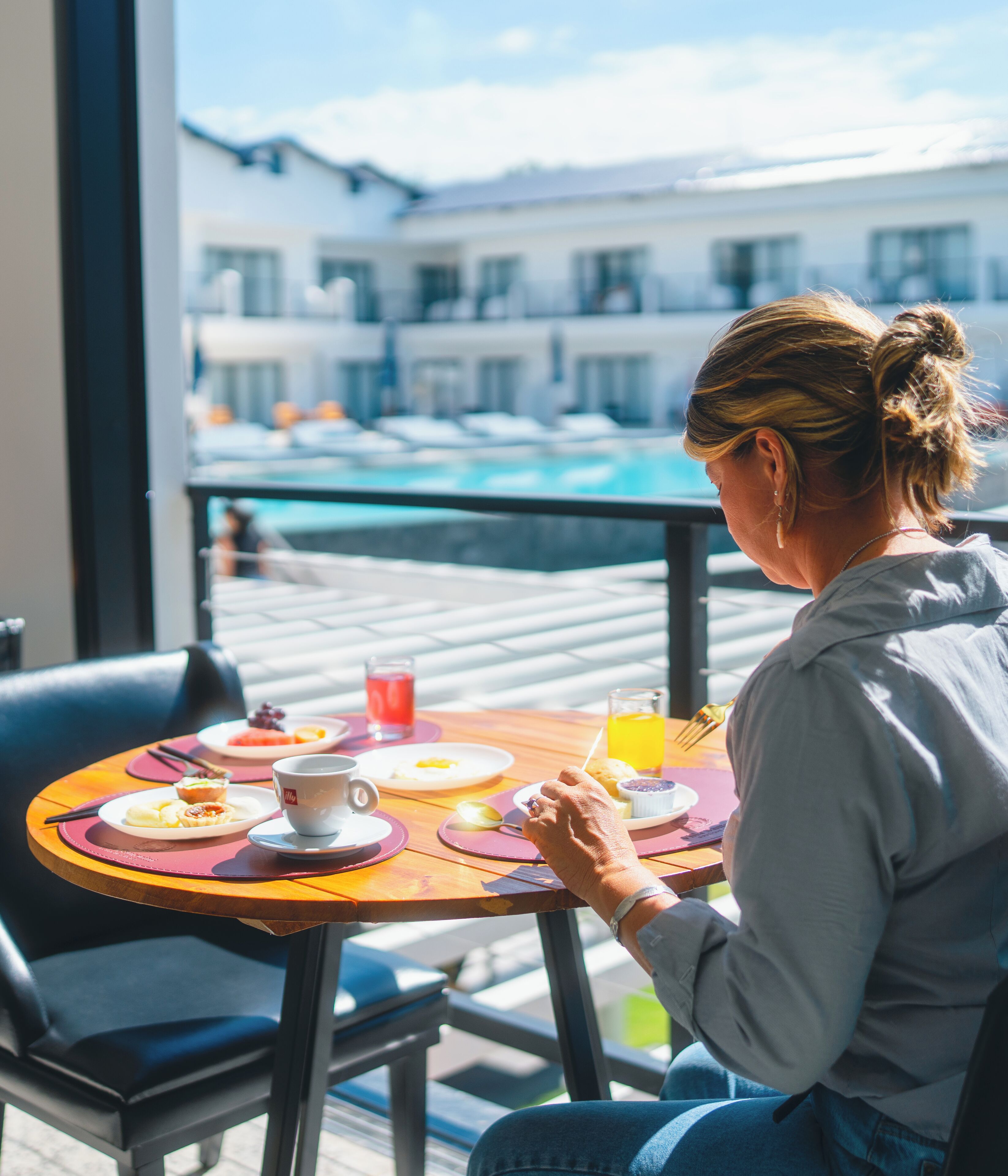 Une personne est assise à une table à l’extérieur, devant un petit-déjeuner, et a devant elle une piscine et des bâtiments.