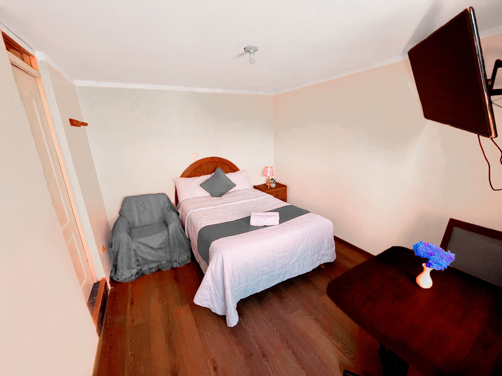 A bedroom with a bed, bedside table, chair, and a TV mounted on the wall.