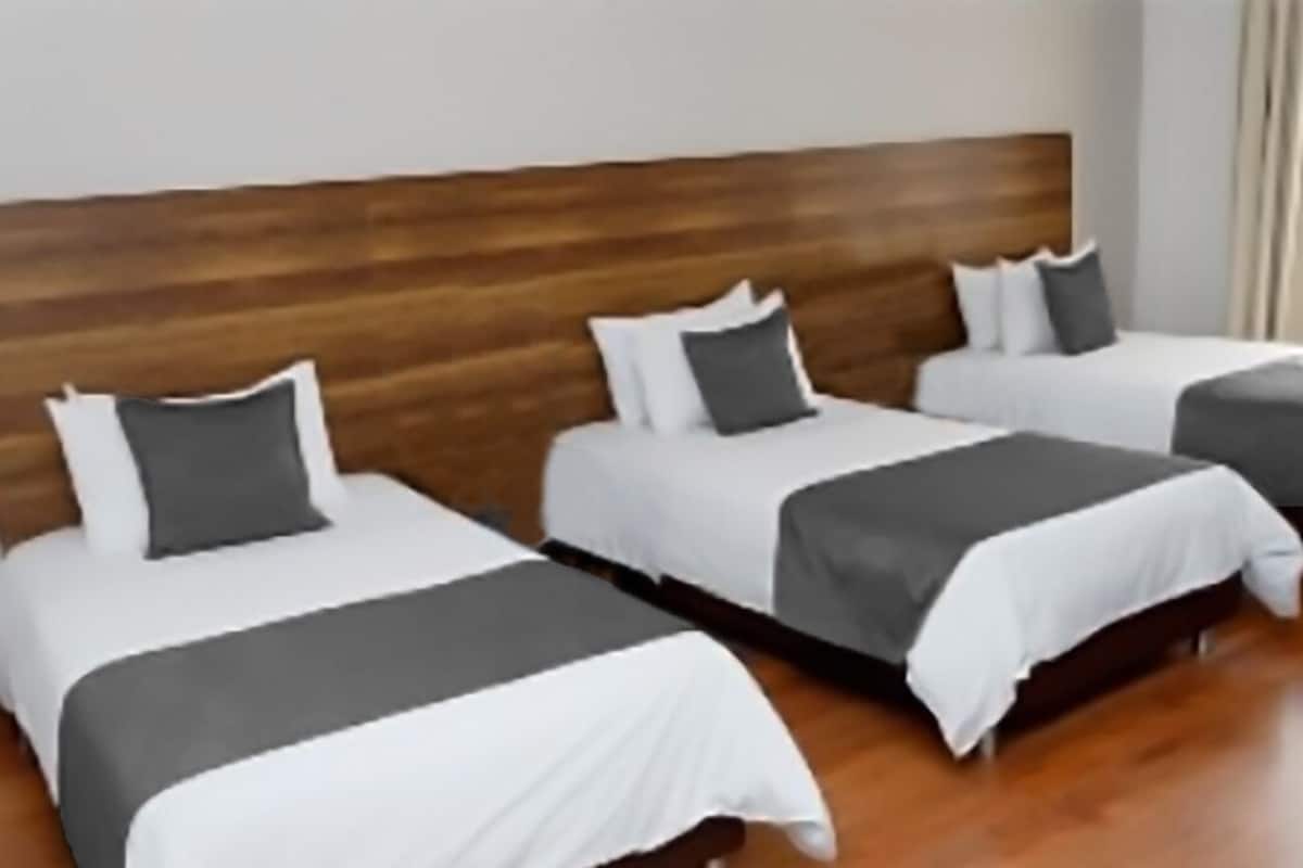 A hotel room with three beds aligned against a wooden headboard, each with white bedding and gray throw pillows.