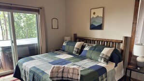 Un dormitorio con una cama grande, cabecera de madera, ventana con vistas a árboles, un cuadro en la pared y una mesita de noche con una lámpara.
