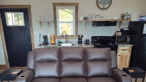 Una sala de estar con un sofá de cuero marrón, una mesa de centro redonda de madera, y una zona de cocina con pileta, estufa y microondas.