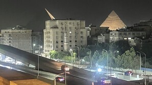 A nighttime cityscape with a prominent pyramid-like structure in the background, modern buildings, and a highway with vehicles. - wake up to the pyramids (Giza)