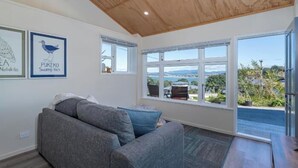 A modern living room with a grey sofa, a wooden ceiling, and large windows offering a view of the outdoors. - Oceanview Tiny Home Whiti (Whitianga)