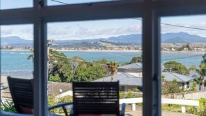 A view from a window overlooking a coastal area with mountains in the distance, a beach, and residential buildings. - Oceanview Tiny Home Whiti (Whitianga)