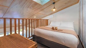 A bedroom with a skylight, a bed, a bedside table, and a wicker chair. - Oceanview Tiny Home Whiti (Whitianga)