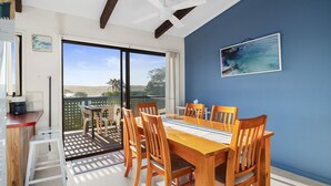 A dining area with a wooden table and chairs, a view of the outdoors, and a painting on the wall. - Pacific Chillz in Ulladulla (Ulladulla)