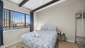 A bedroom with a bed, a window with curtains, a bedside table, and a metal shelf unit. - Pacific Chillz in Ulladulla (Ulladulla)