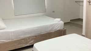 A room with two beds, one with a headboard and the other without. There is a window with blinds, a door, and a small shelf.