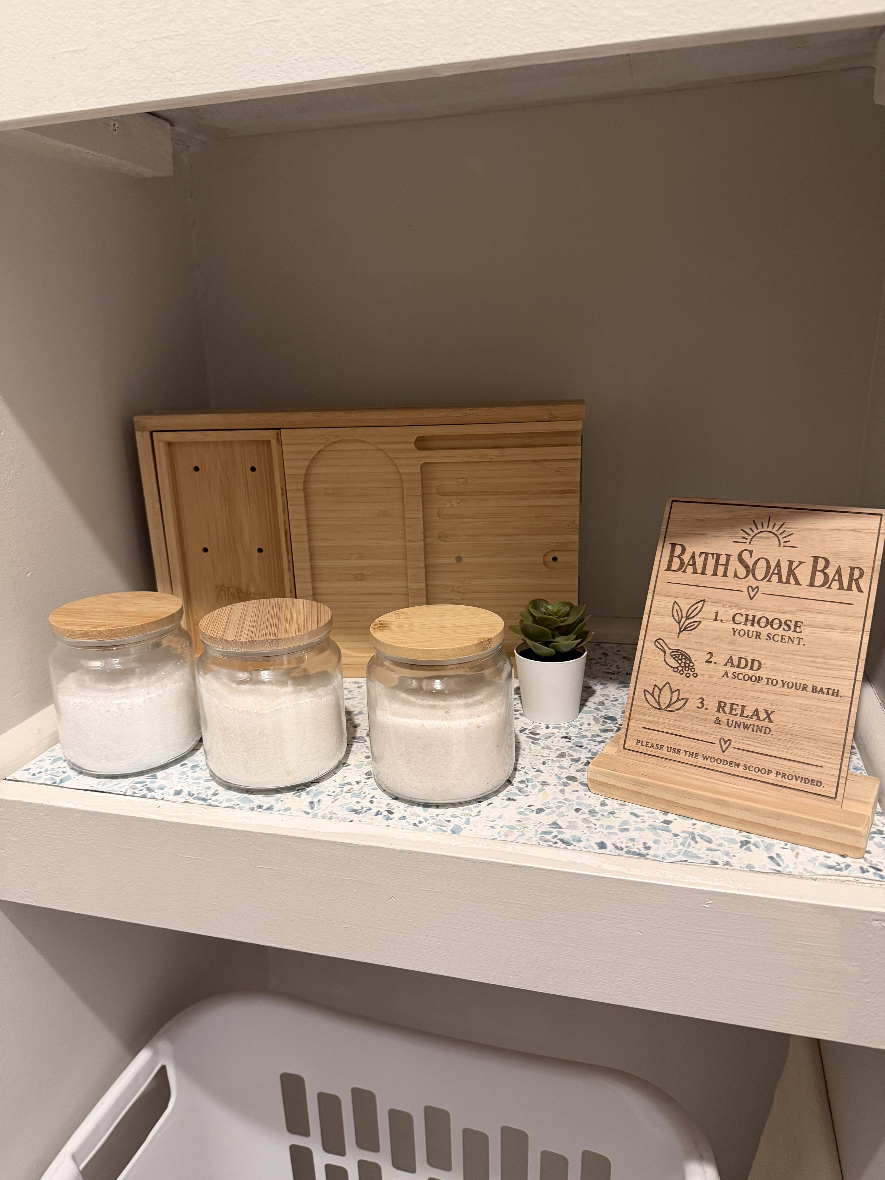 Ein Badezimmer-Waschtisch mit einem Holzkabinett, drei Glasbehälter mit Deckeln, einer kleinen Topfpflanze und einem Holzschild mit Anweisungen für eine Badesalzen-Anwendung.