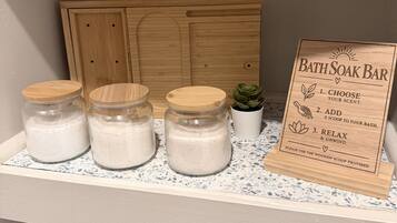 Ein Badezimmer-Waschtisch mit einem Holzkabinett, drei Glasbehälter mit Deckeln, einer kleinen Topfpflanze und einem Holzschild mit Anweisungen für eine Badesalzen-Anwendung.