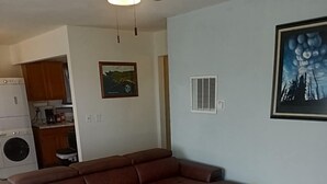 A living room with a sectional sofa, a patterned rug, a ceiling fan, a kitchen area, and a framed picture on the wall.