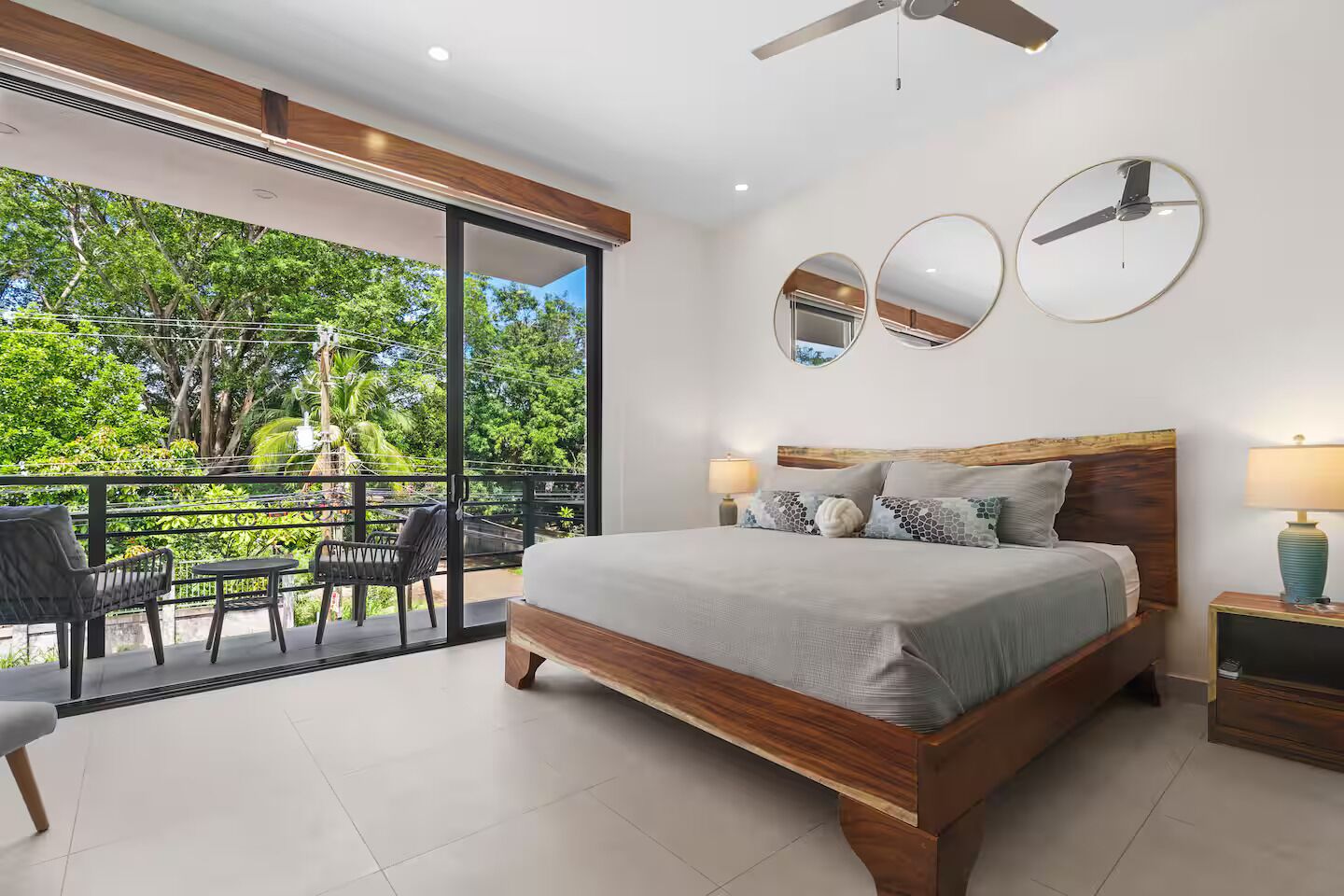 A bedroom with a large bed, two bedside tables, a ceiling fan, and a balcony with outdoor furniture.