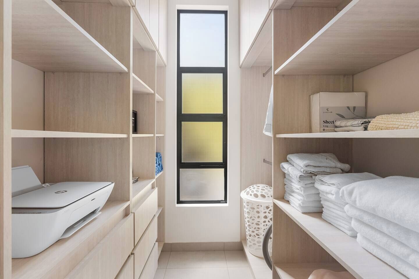 A modern, narrow walk-in closet with wooden shelving, a laptop on the left, neatly folded towels on the right, and a window with frosted glass panels.