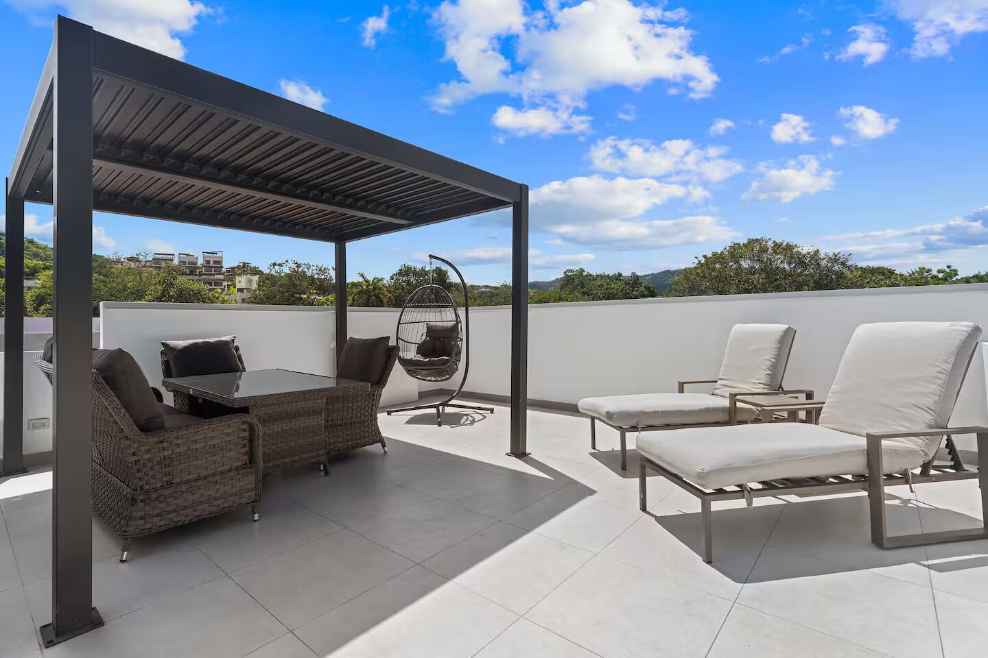 A rooftop patio with a pergola, wicker furniture, a hanging chair, and white lounge chairs.