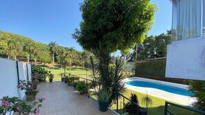 A pool area with a tiled pathway, potted plants, and a view of a grassy field and trees. - Apartment with ocean and golf course views (Acapulco de Juárez)